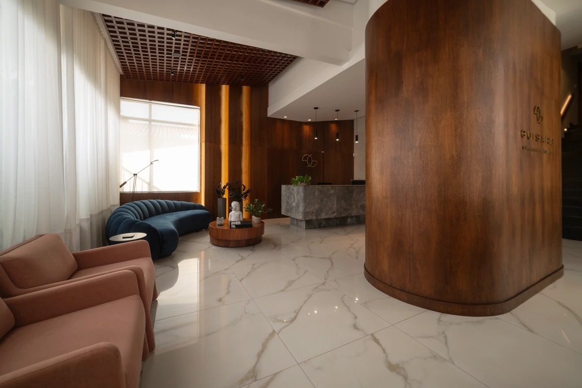 Recepção da clínica, com sofá curvo azul, piso de mármore e painel curvo de madeira